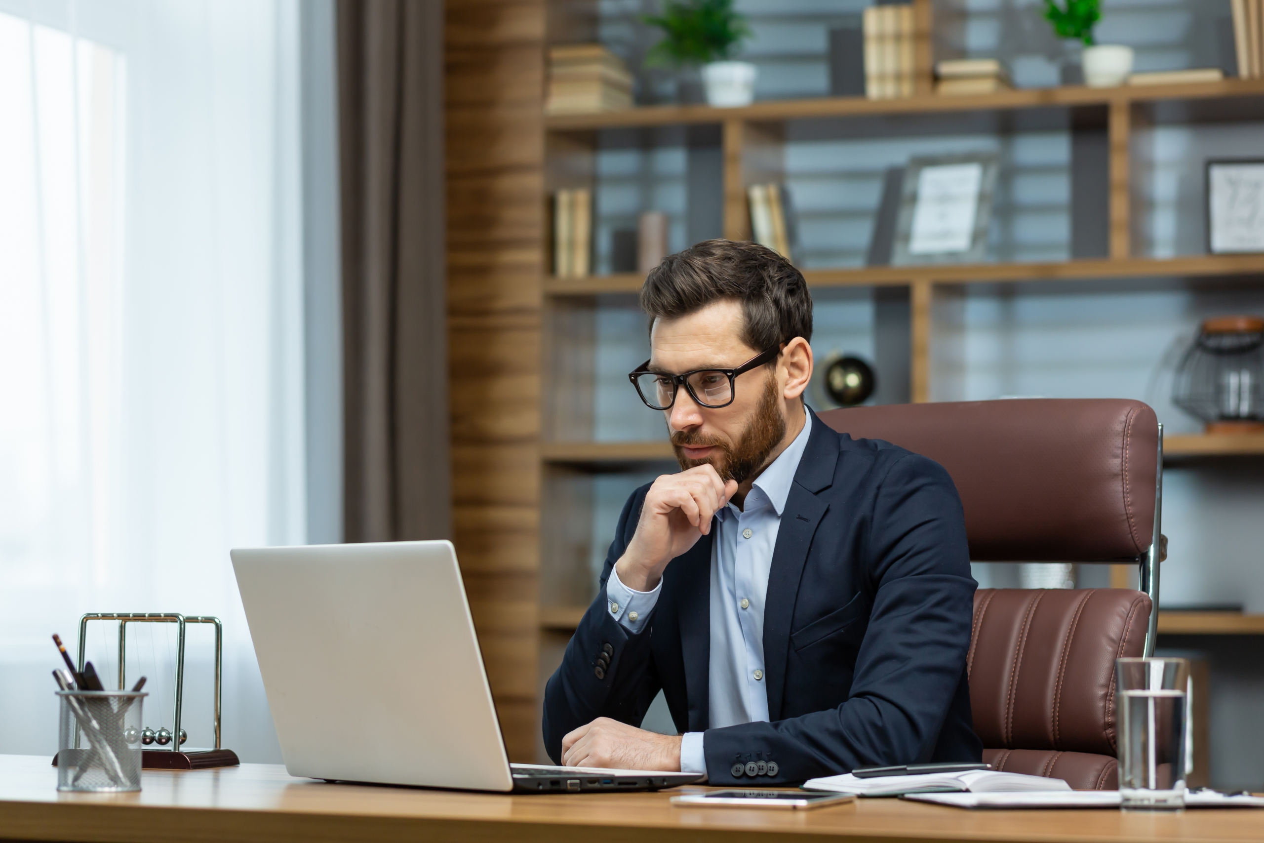 Thinking businessman working sitting at desk, mature adult boss in business suit and beard looking at laptop screen thinking about financial investment decisions inside office